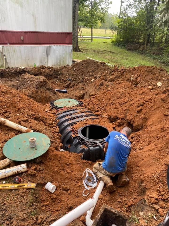 Man installing septic system in an excavated area outdoors, next to a building and lawn.