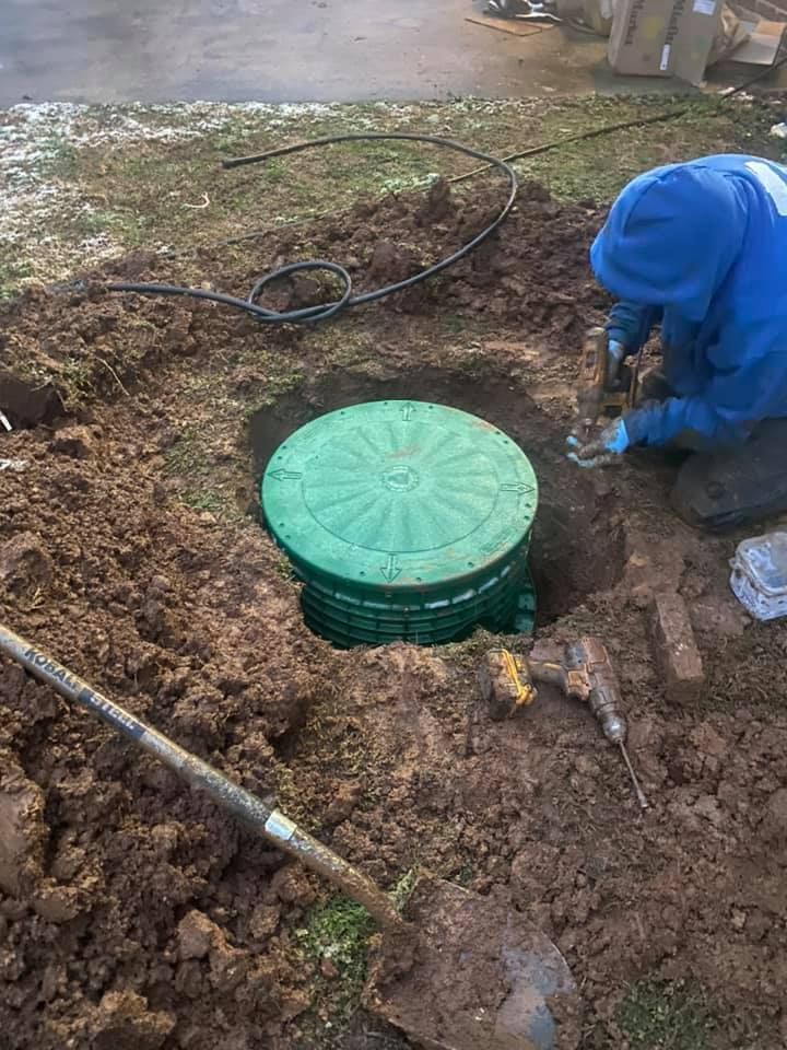 A person in blue jacket works on installing a green septic tank lid in a muddy hole outdoors.