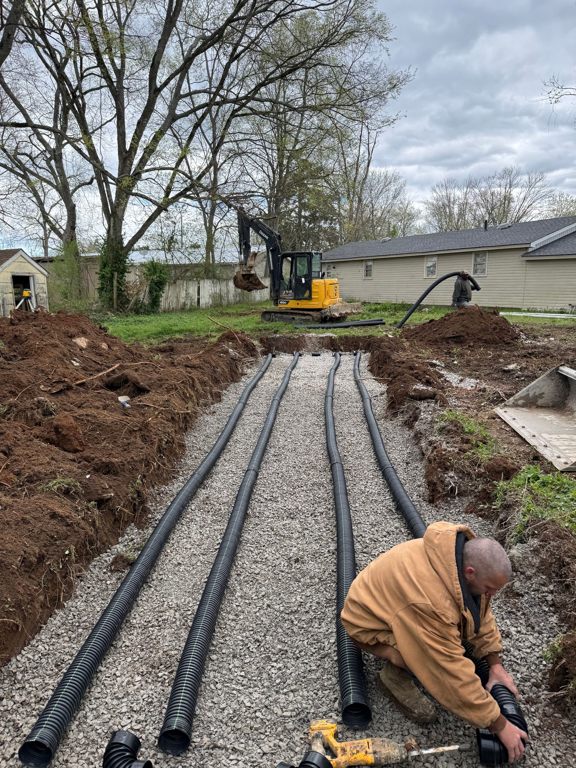 Man connects pipe in a trench with gravel, excavator in the background, cloudy sky.