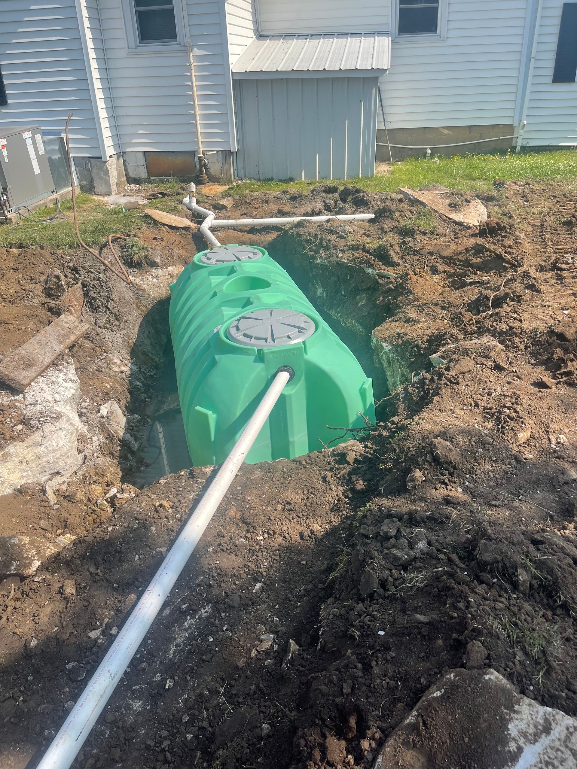 Green septic tank in a hole, connected to white pipes, near a house.
