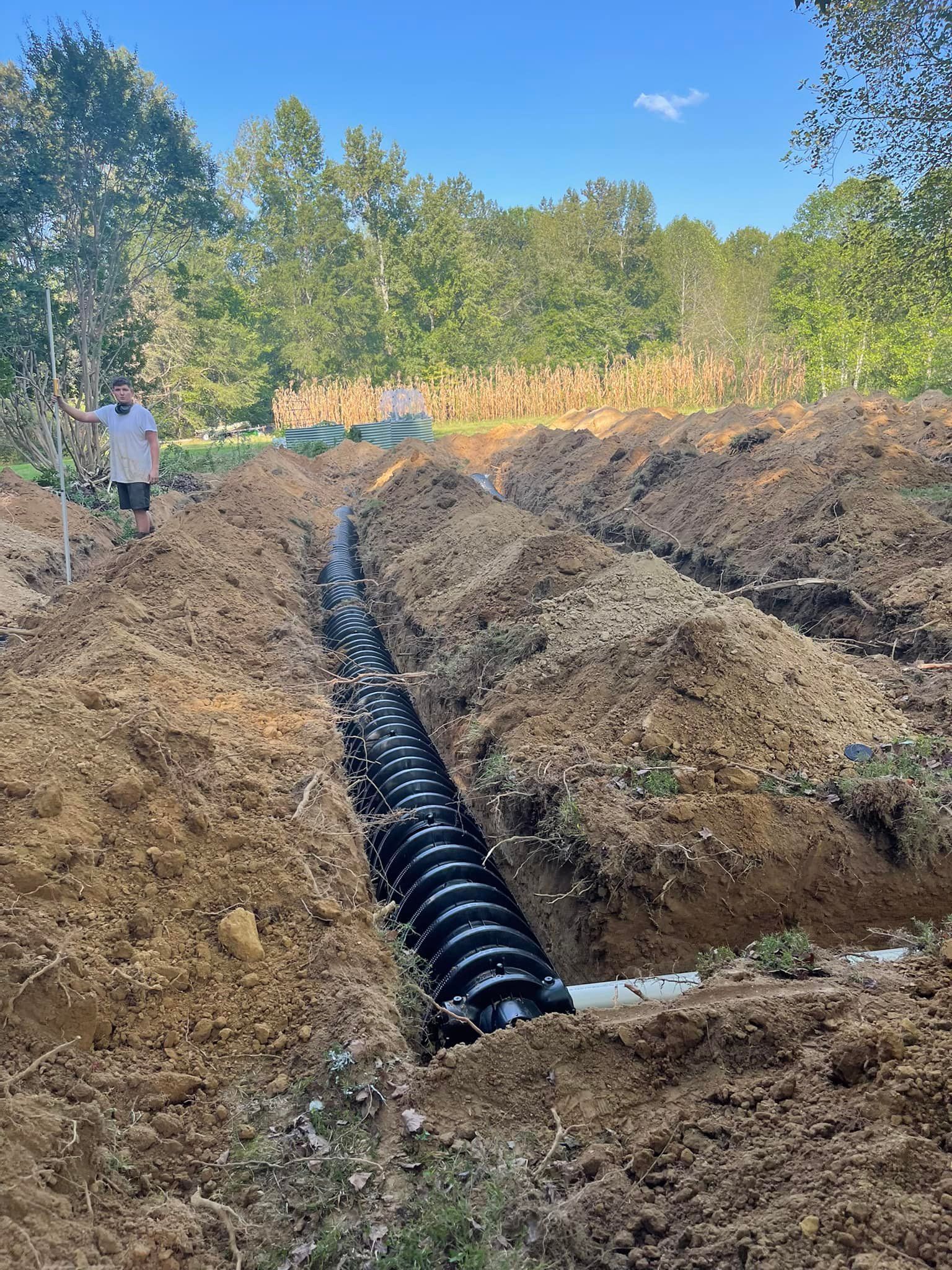 A trench with black drainage pipe and a person standing in a rural, outdoor setting.