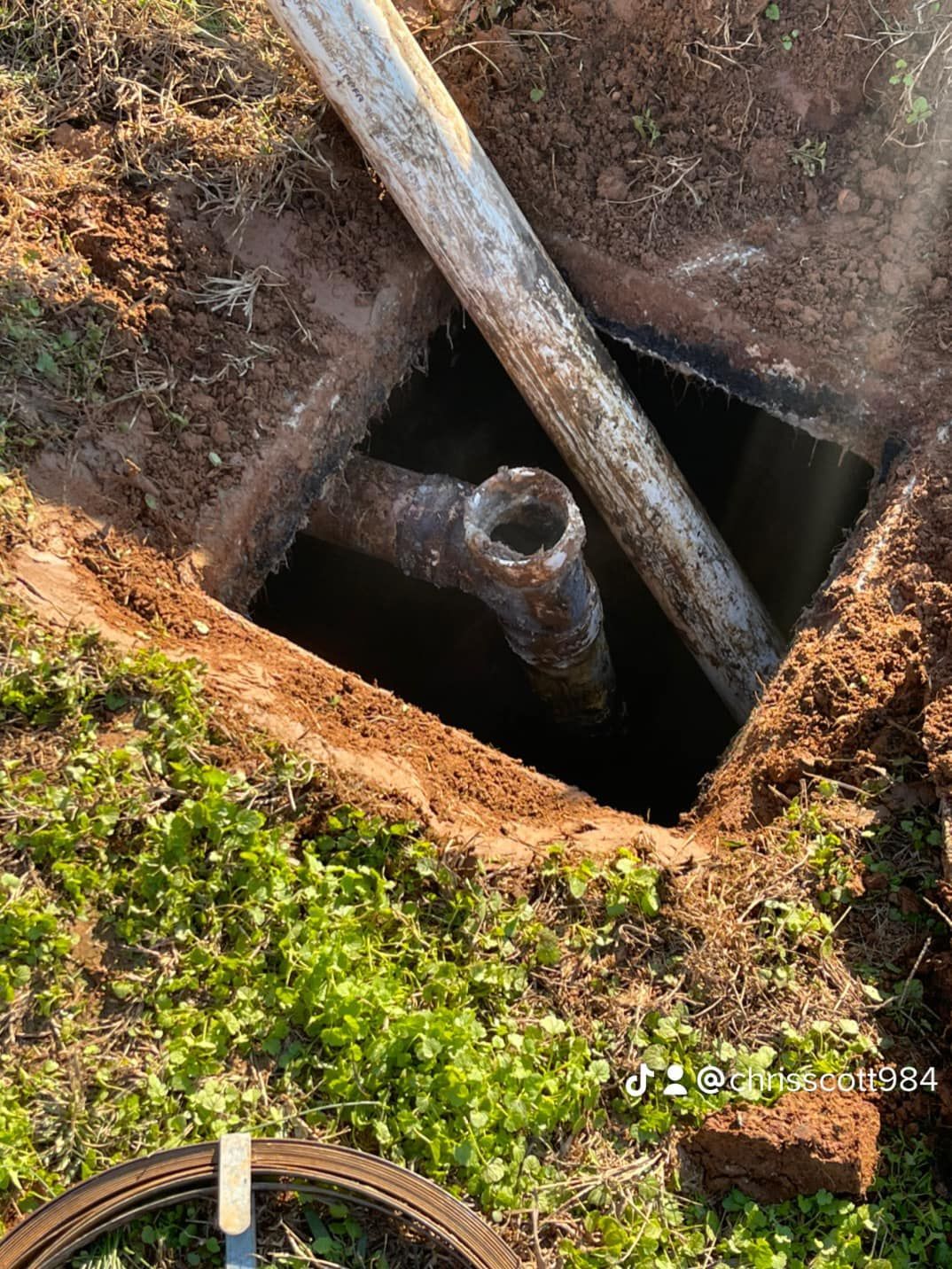 A square hole dug in the ground reveals a rusty pipe and a white pipe. Dirt and grass surround the hole.