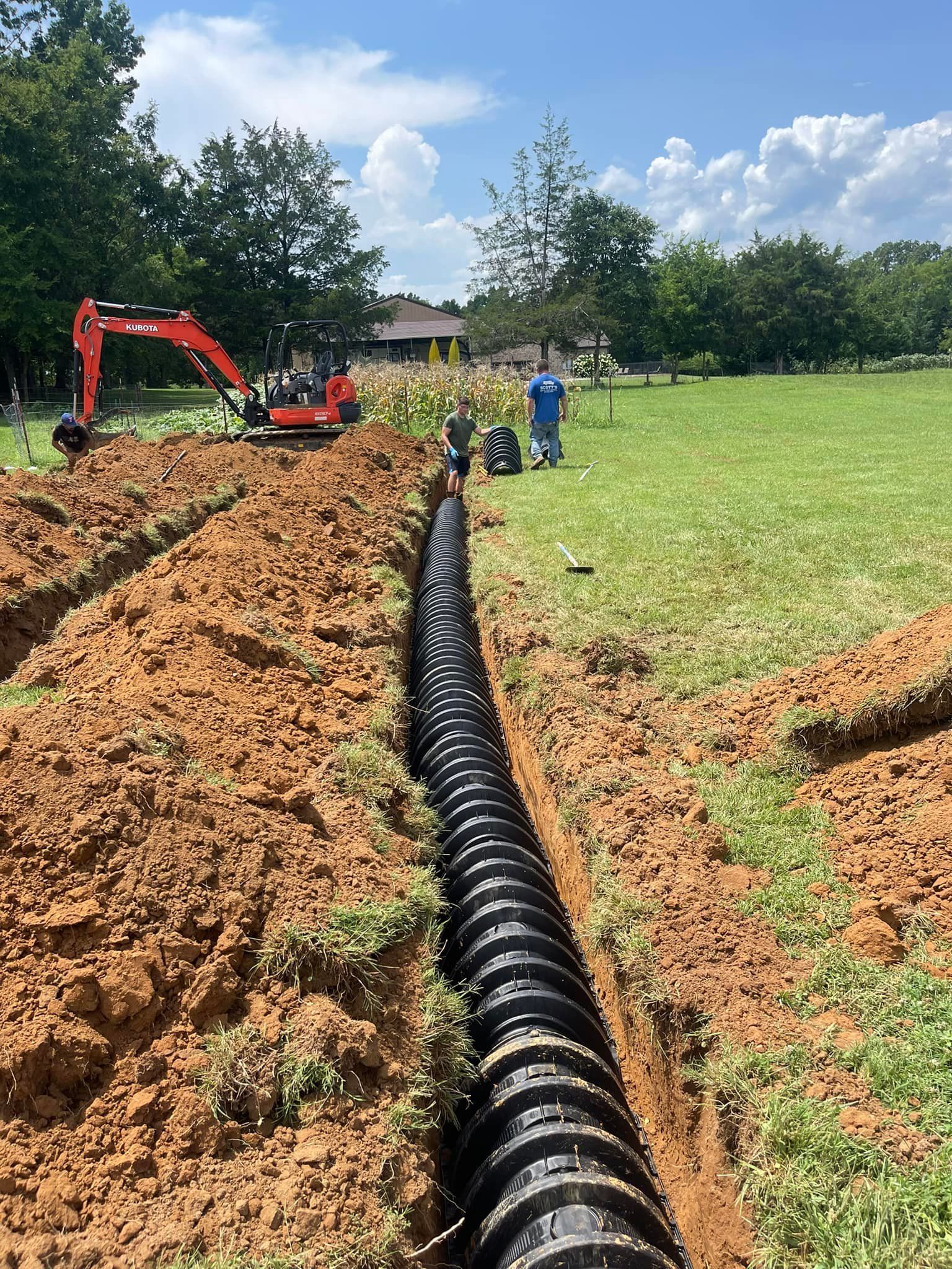 Trench in grassy area with black drainage pipe, excavator, and workers under blue sky.