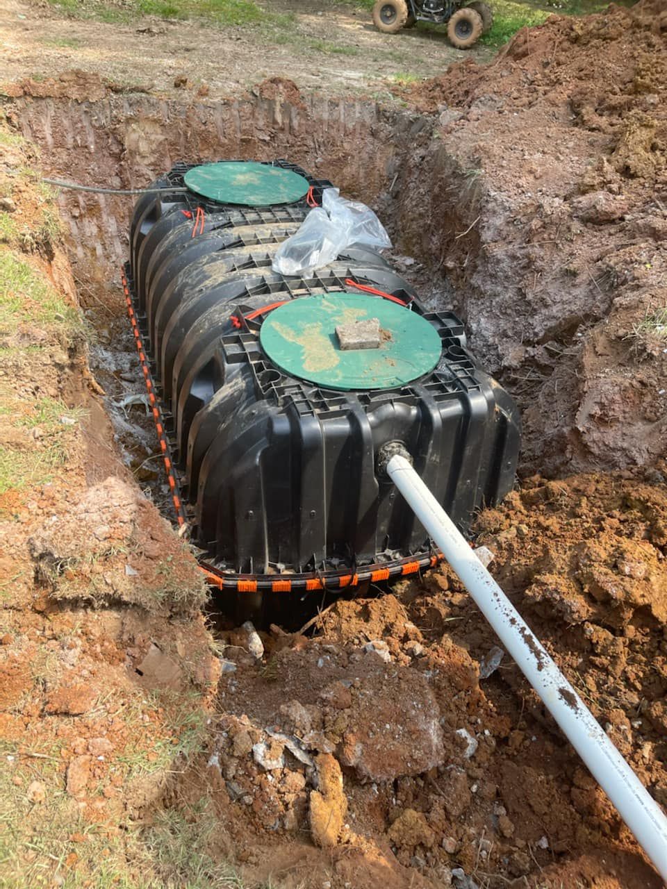 Black septic tank in an excavated trench, with two green access covers and white outflow pipe.
