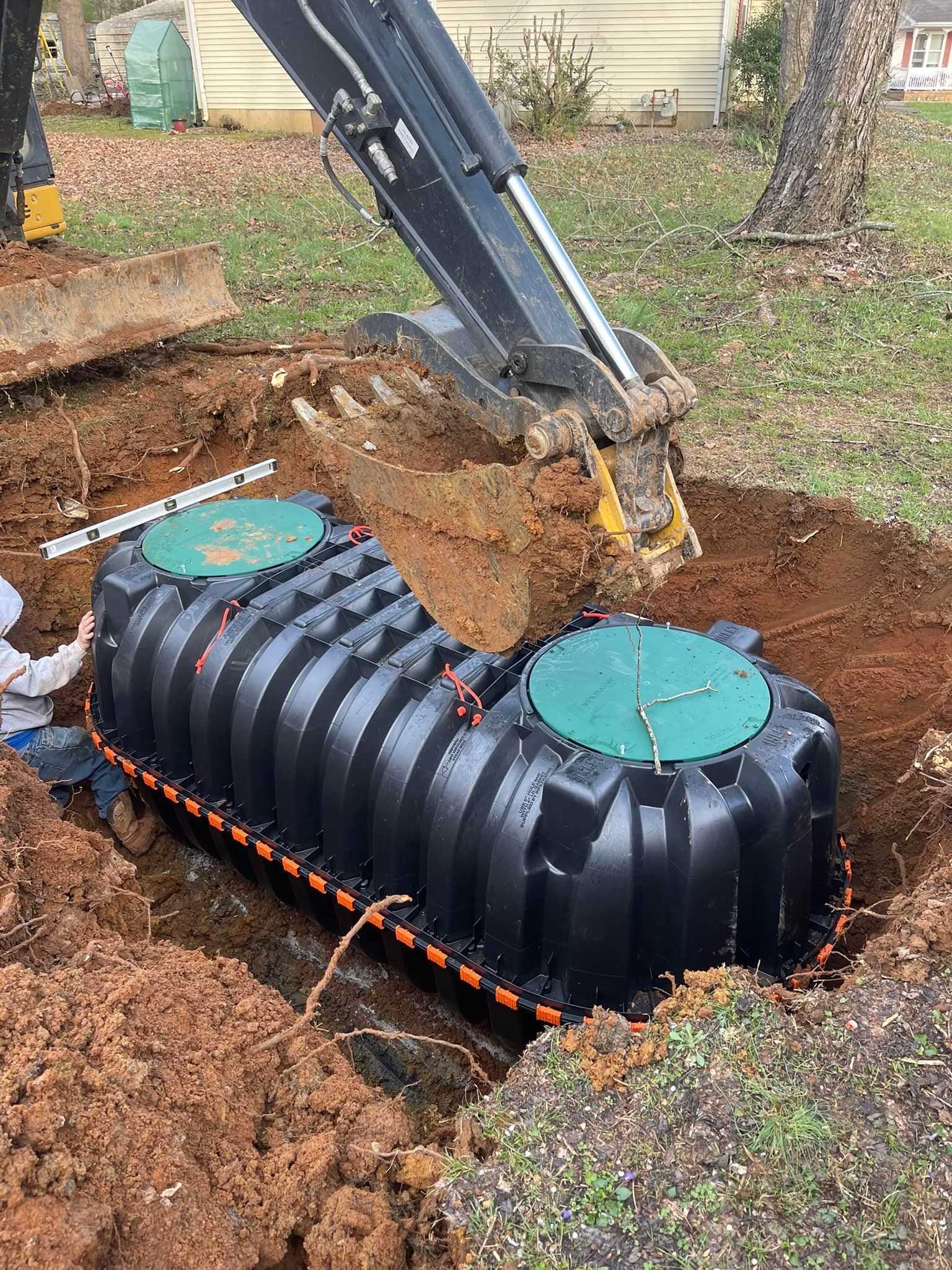 Black septic tank being installed in a hole with an excavator. Dirt and grass surround it.