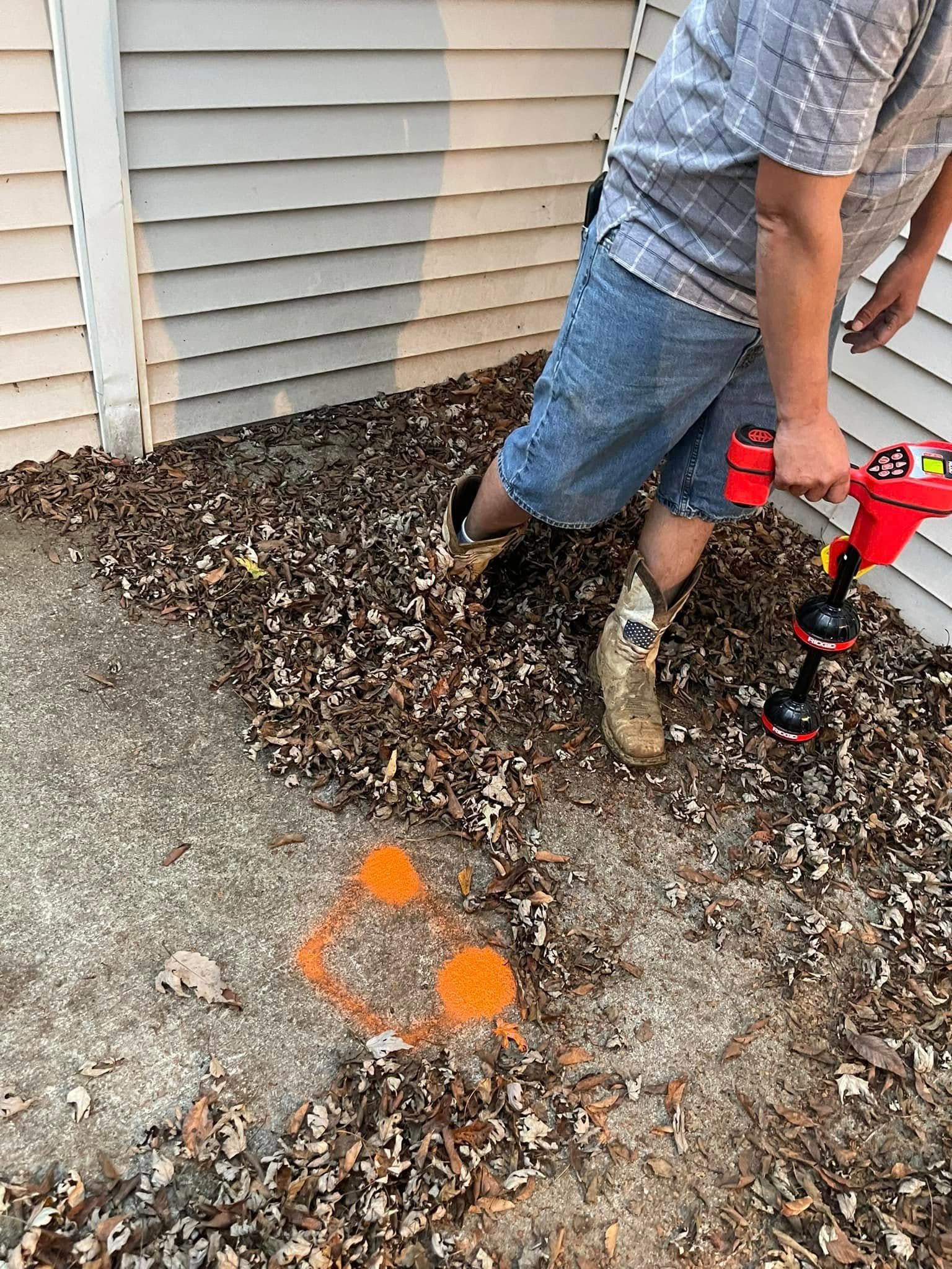 Person uses a detector near orange markings on a concrete surface, beside a building. Brown leaves cover the ground.