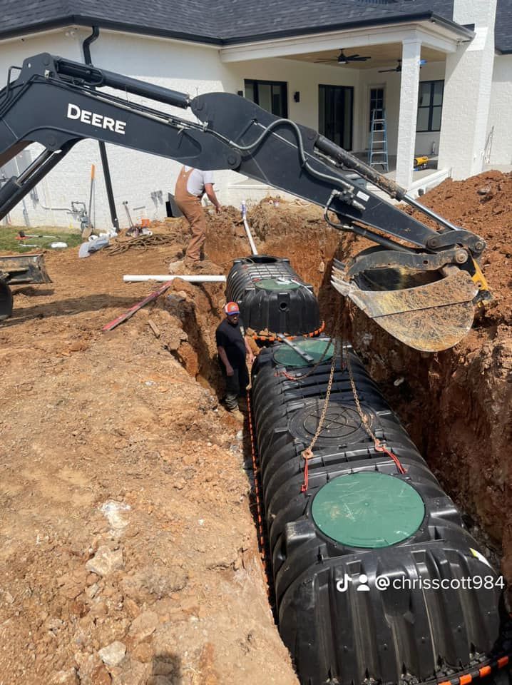 A backhoe lowers a septic tank into a trench, with a worker assisting. Residential construction site.