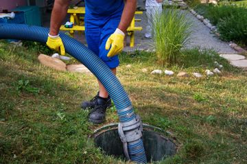 A worker in blue overalls and yellow gloves holding a hose over a septic tank.