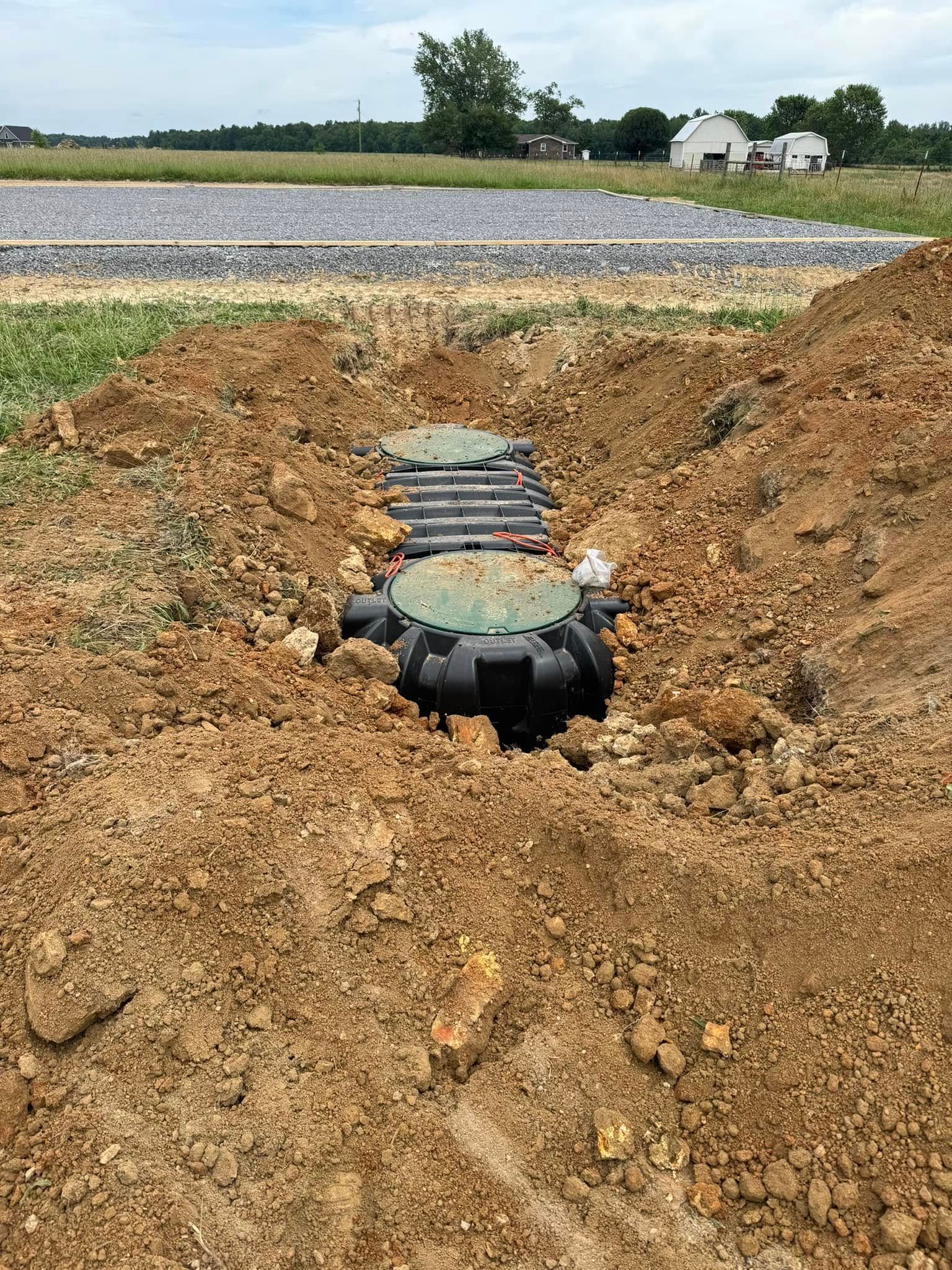 A septic system installation in progress. Black tanks are partially buried in brown dirt, with green access lids visible.