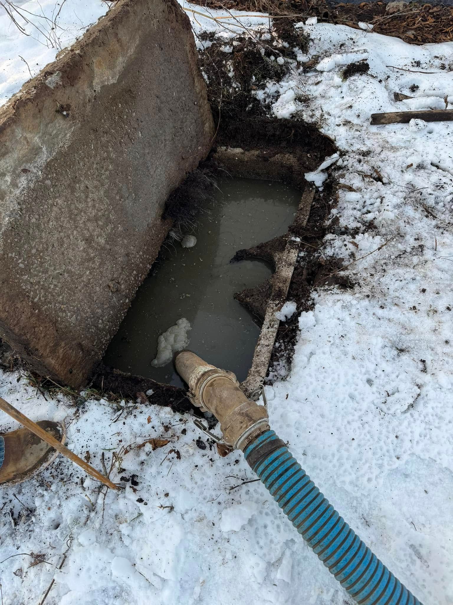 Septic tank with open lid, water, and blue hose surrounded by snow.