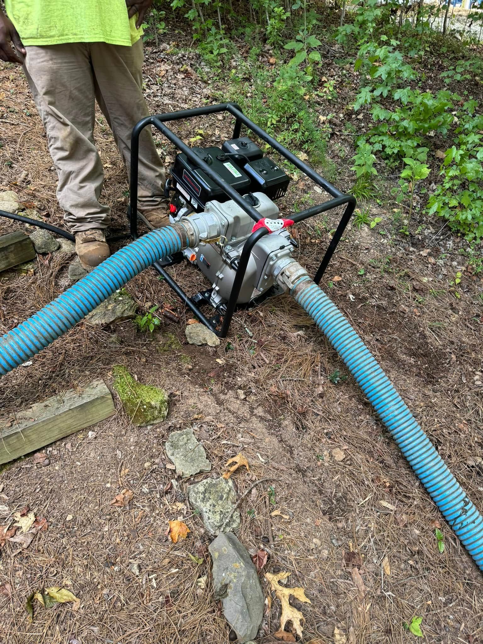 Person operating a water pump with blue hoses in a wooded area.