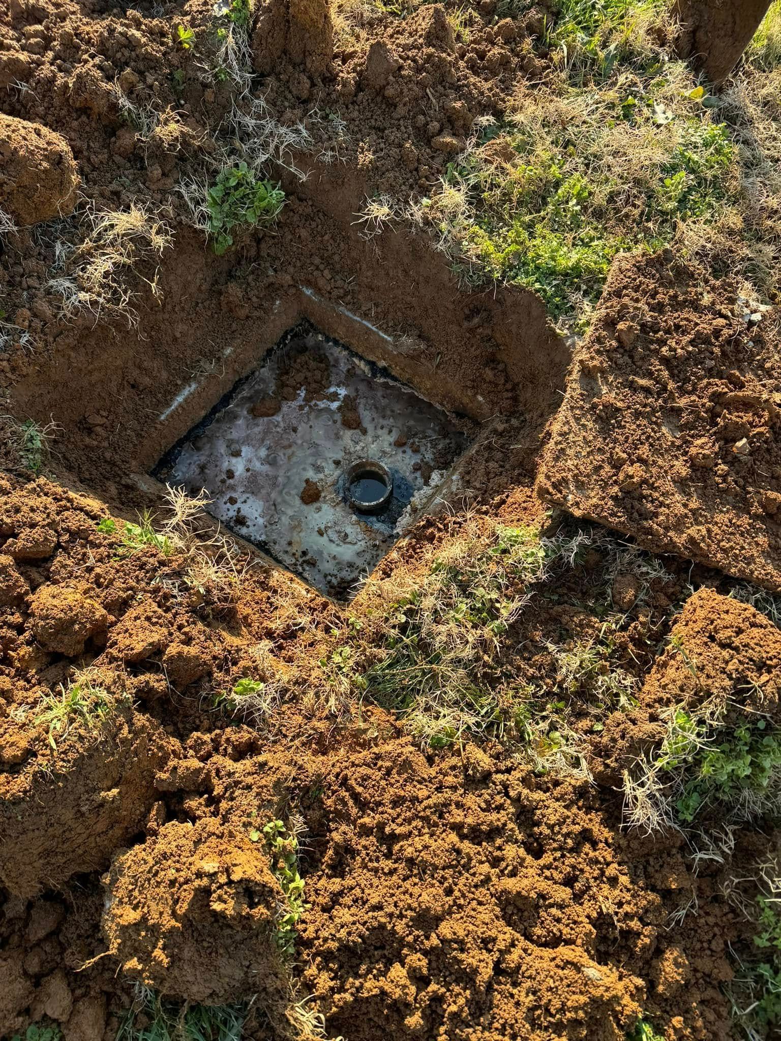 Square metal utility access cover in the ground, surrounded by brown soil and grass.