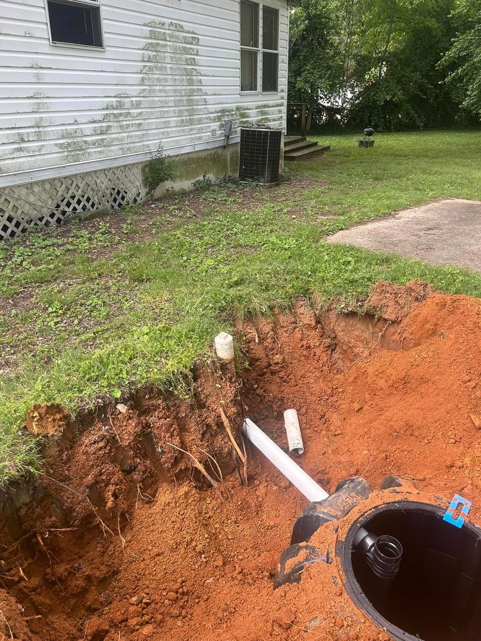 Excavated area near a house, revealing pipes and a dark tank. Red soil and green grass surround.