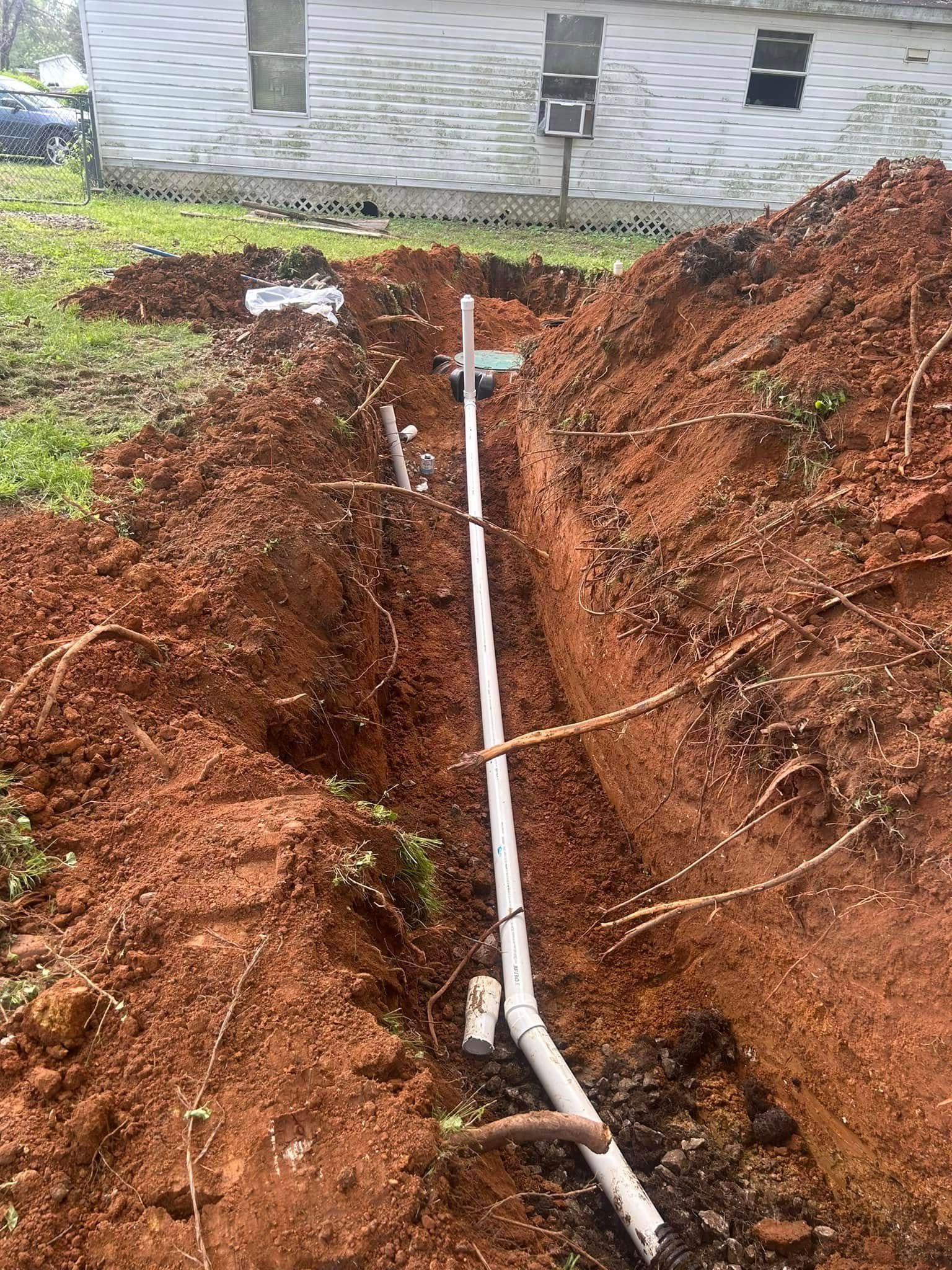 Trench dug in red soil, holding white PVC pipe, near a house with grass and trees.