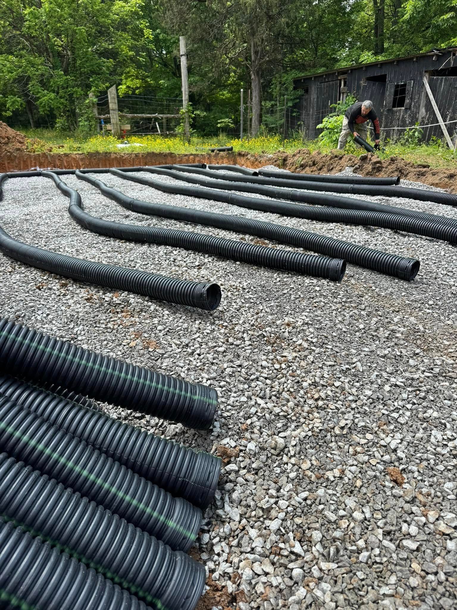 Black corrugated pipes laid over gravel, likely for drainage; a person works on a shed in the background.