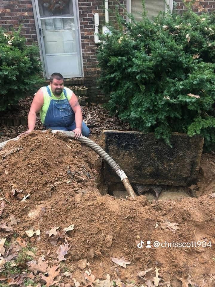 Man kneeling by an open septic tank, holding a hose. Earth is piled around the tank.
