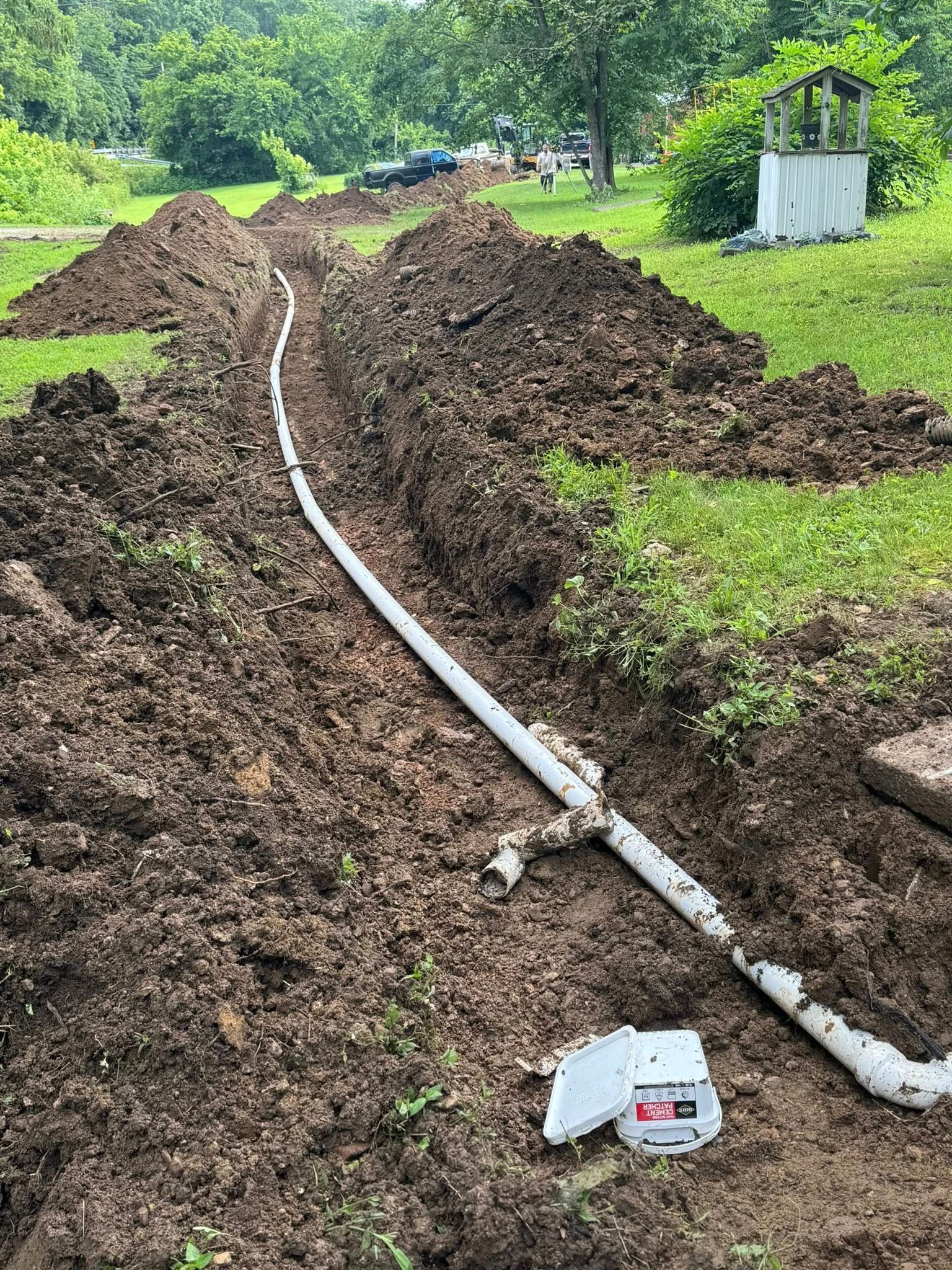 Long trench with white pipe and dirt mounds in a grassy area.