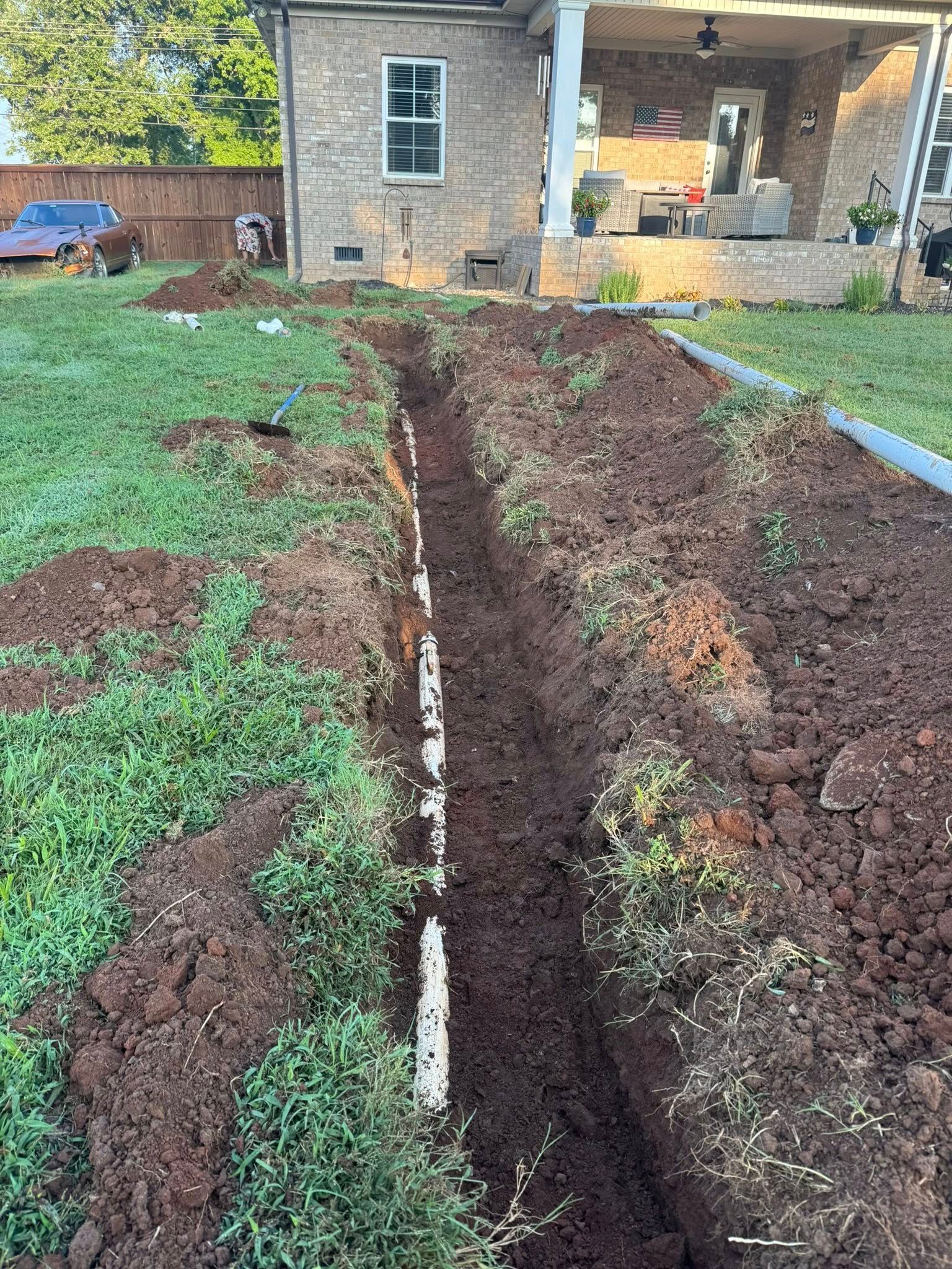 Trench in a yard, showing exposed white pipe, leading towards a brick house with a covered patio.