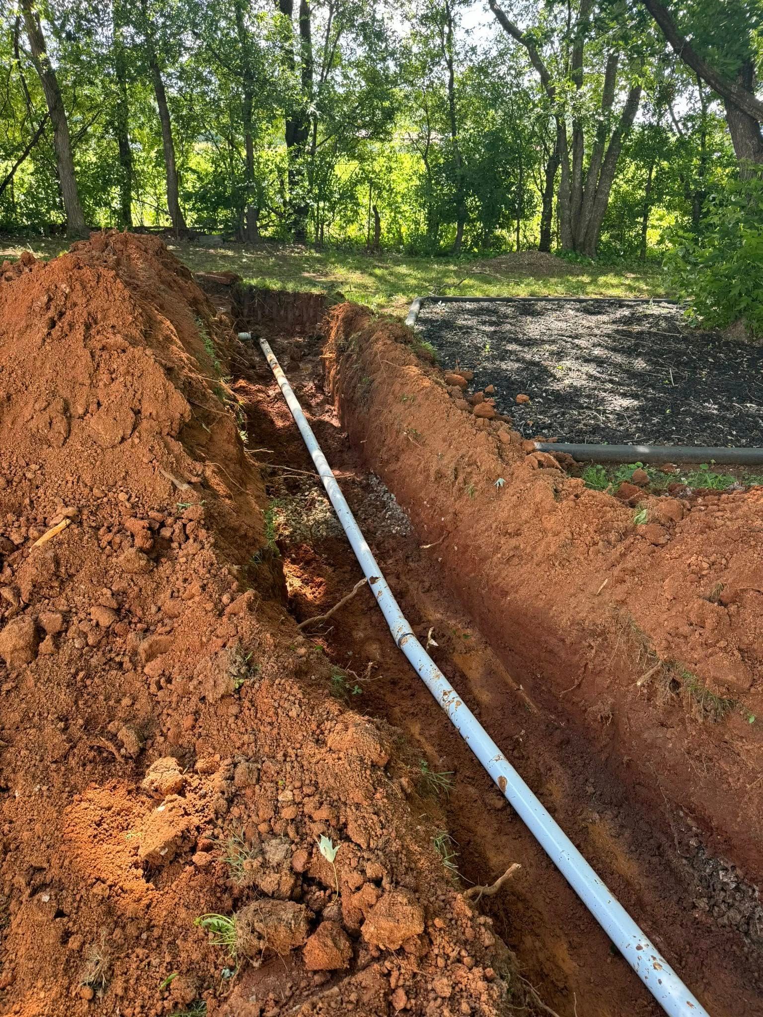 Trench dug in red soil, holding a white pipe. Green trees in the background.