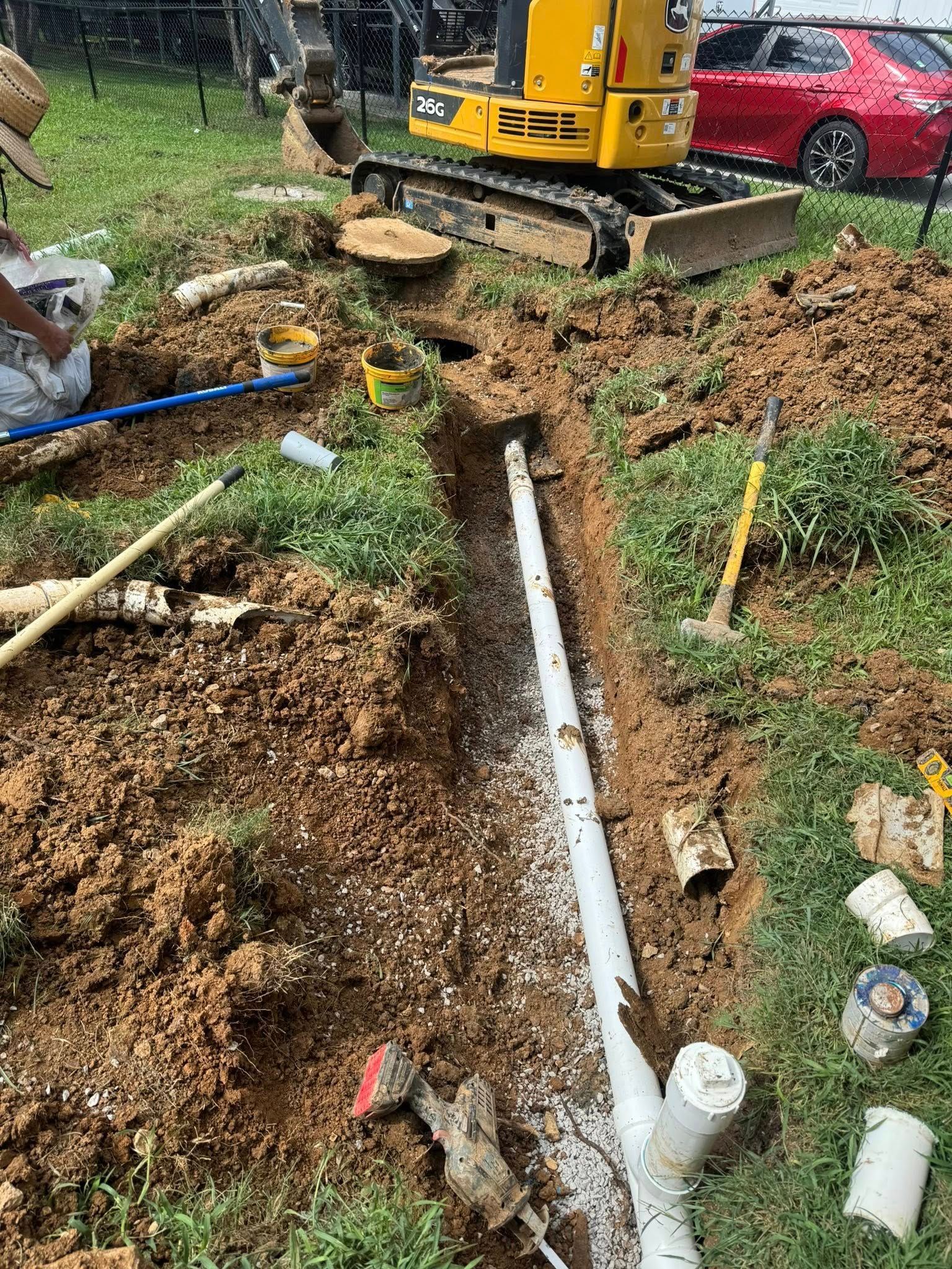 A backhoe digs a trench for PVC pipes in a grassy area, a red car is parked in the background.