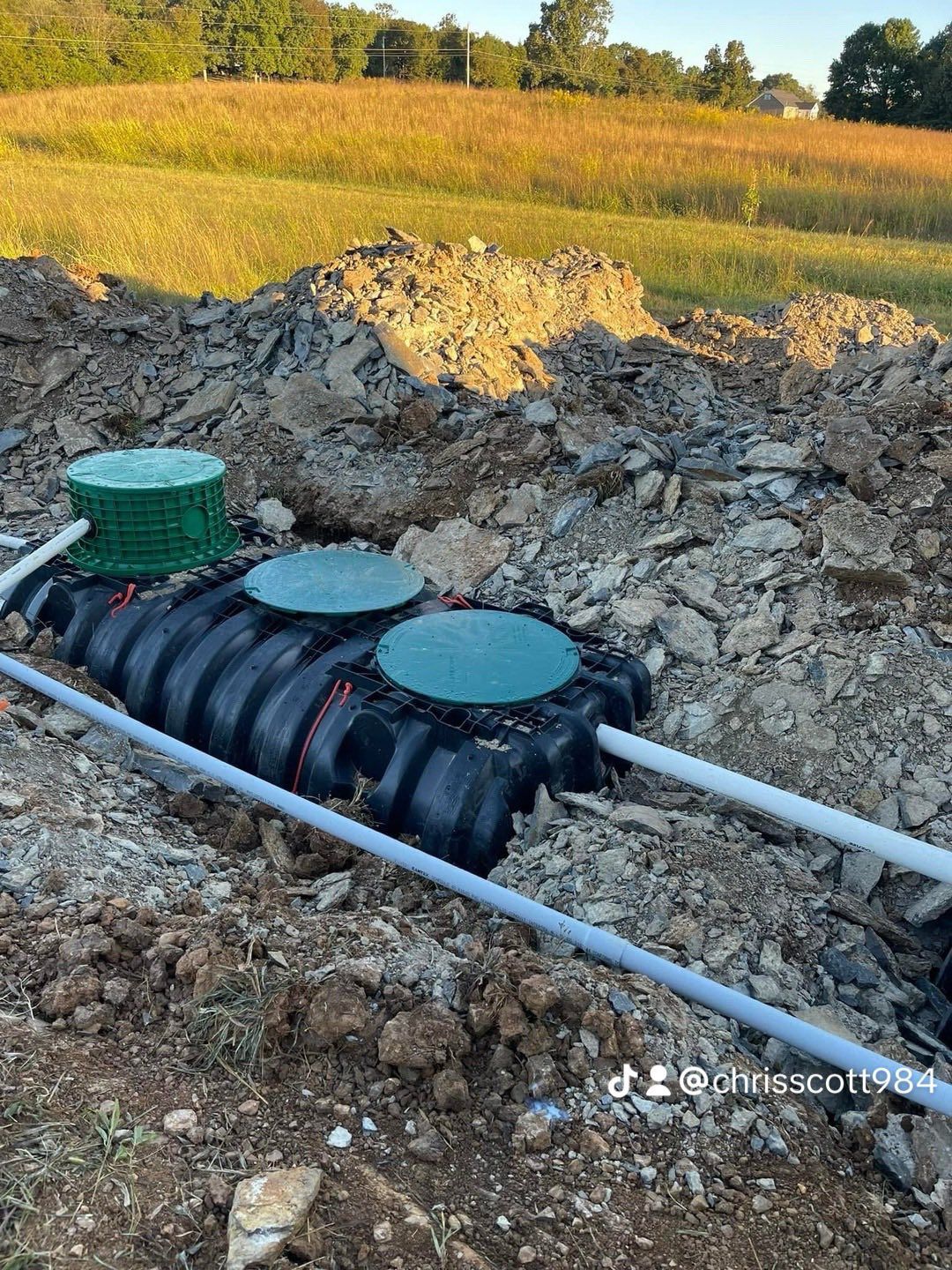 Septic tank installation in a field: black tank, green lids, white pipes, and dirt.