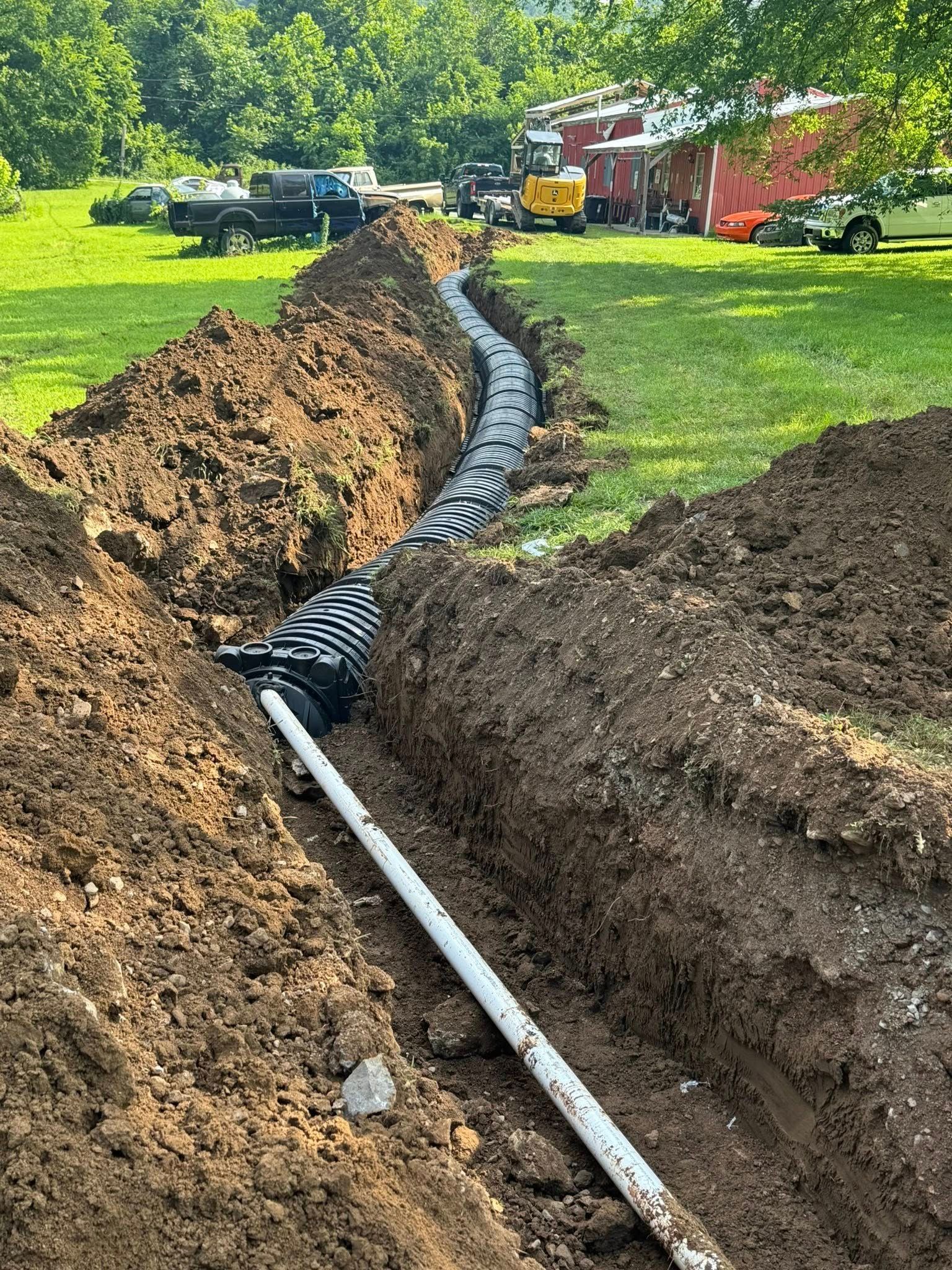 Trench in a grassy area with drainage pipes installed. A truck and equipment are in the background.