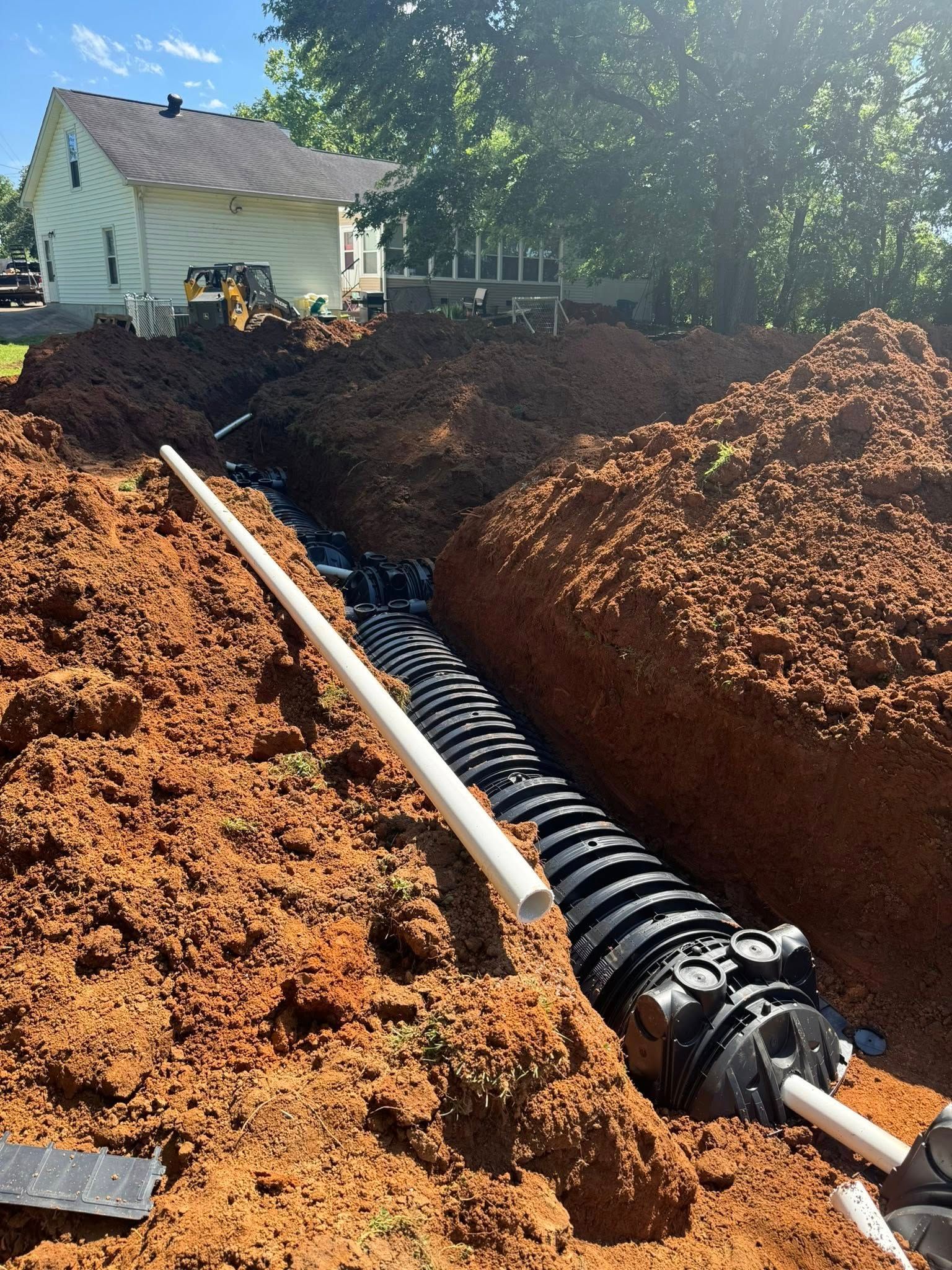 Trench with black plastic drainage system being installed in a yard, with a white house in the background.
