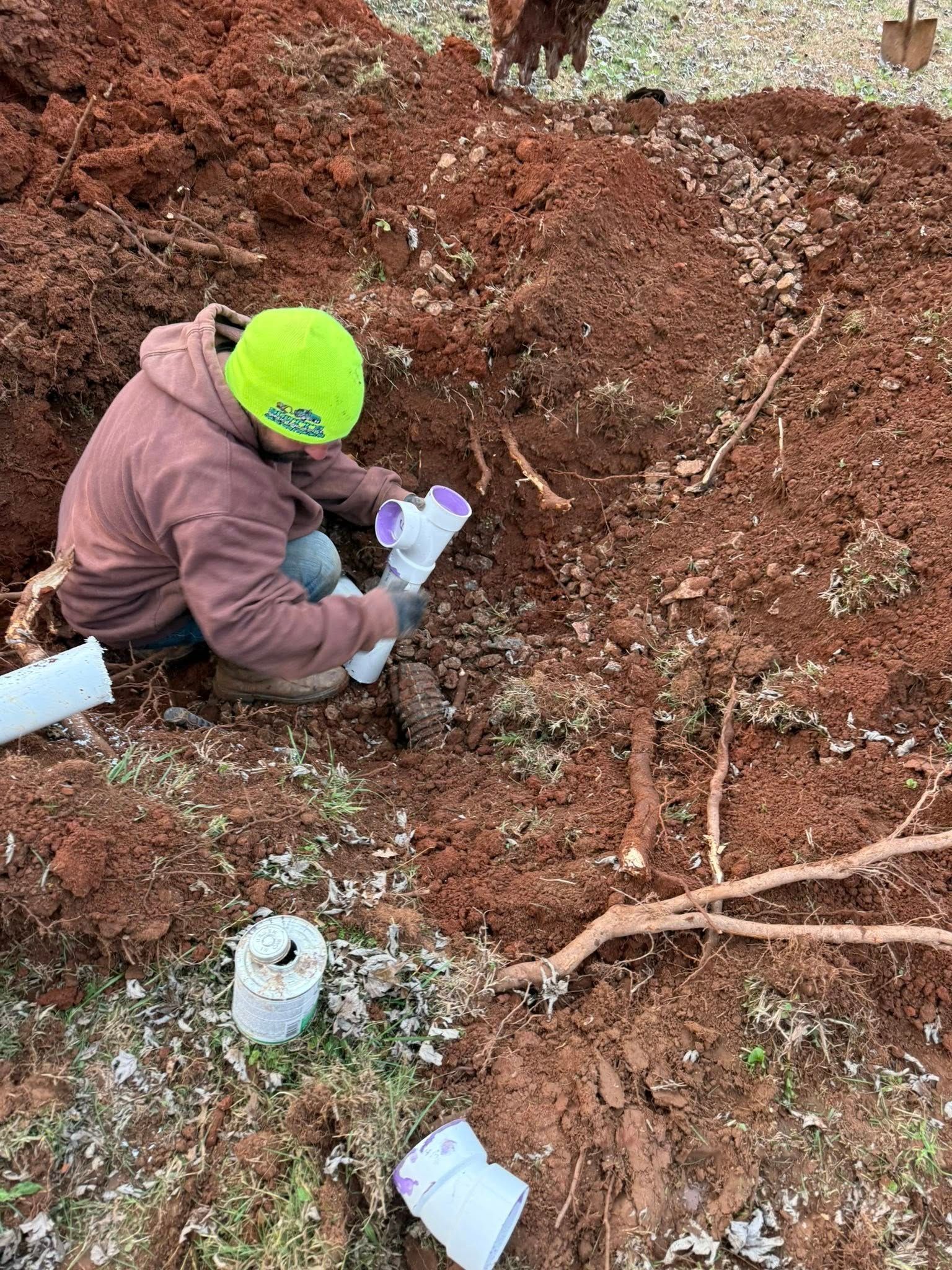 Person in a hard hat working on plumbing in a muddy trench.