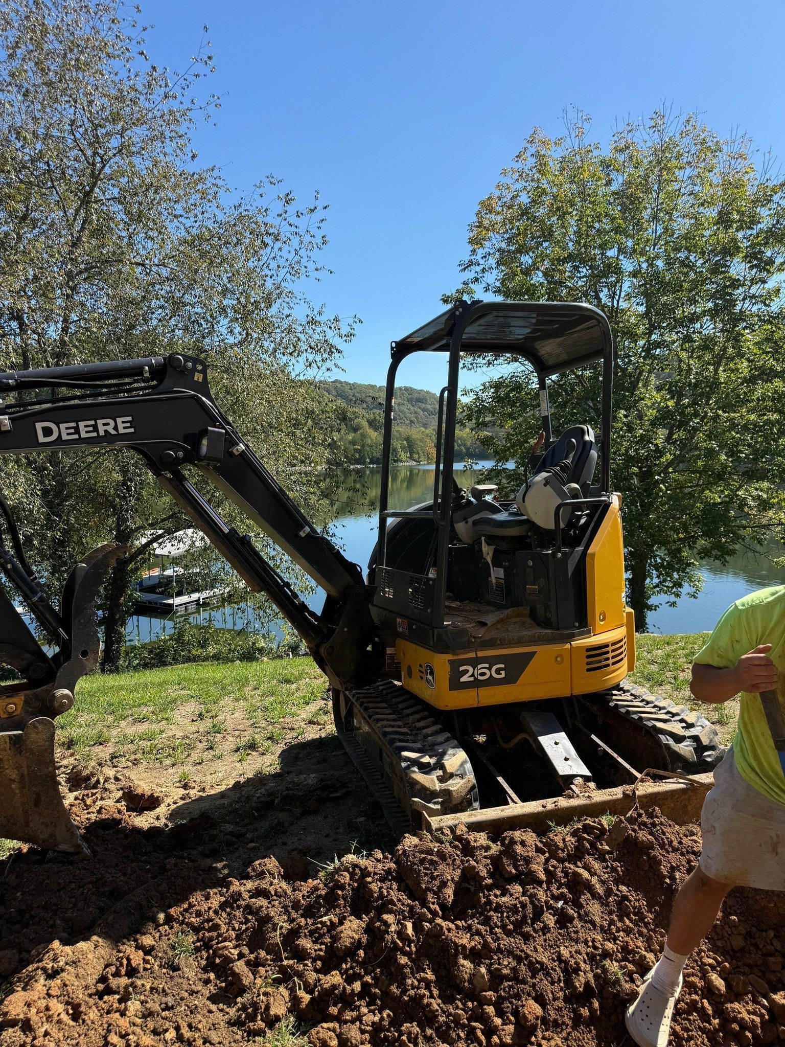 Yellow John Deere mini-excavator digging a trench on a sunny day near water. A person stands nearby.