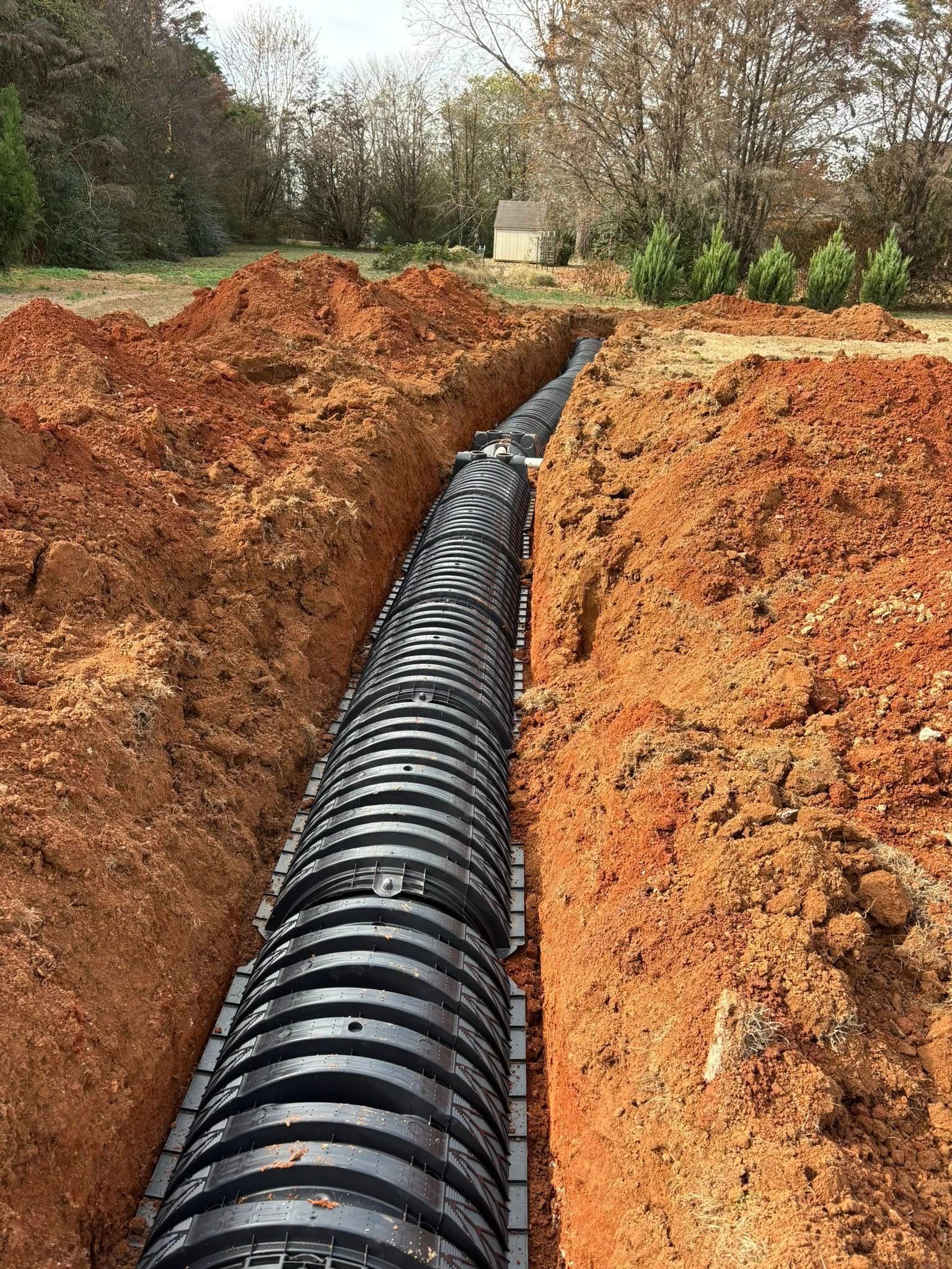 Trench with black corrugated drainage pipe installed, surrounded by red soil, ready for burial.