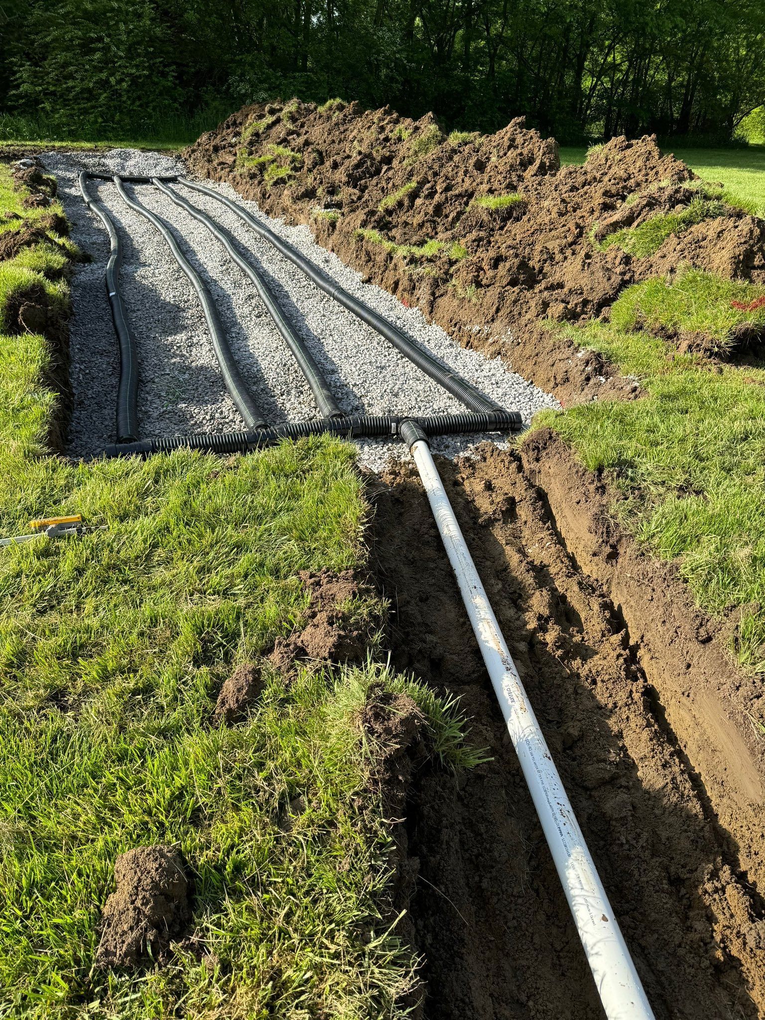 Trench with gravel and pipes laid for drainage system. White PVC pipe connects to the buried system.