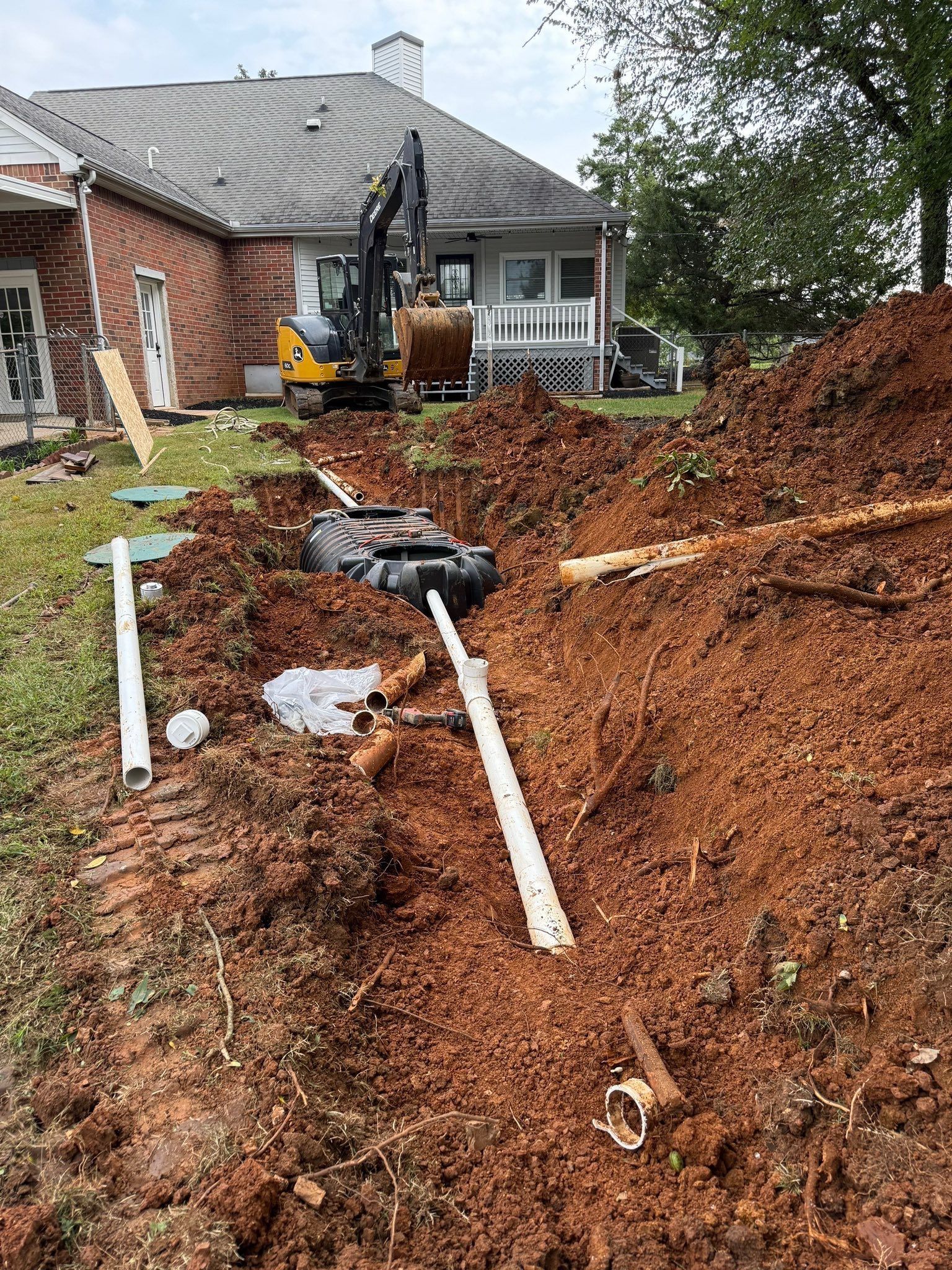 Excavation work in progress near a house. A backhoe and trenches filled with dirt and pipes.