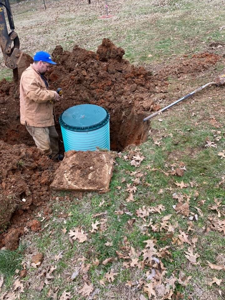 Man working on a buried septic tank. He is in a hole, surrounded by dirt and fallen leaves.