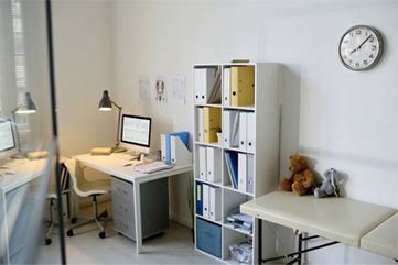 Bright home office with white desk, shelving, lamp, computer, chair, and wall clock.