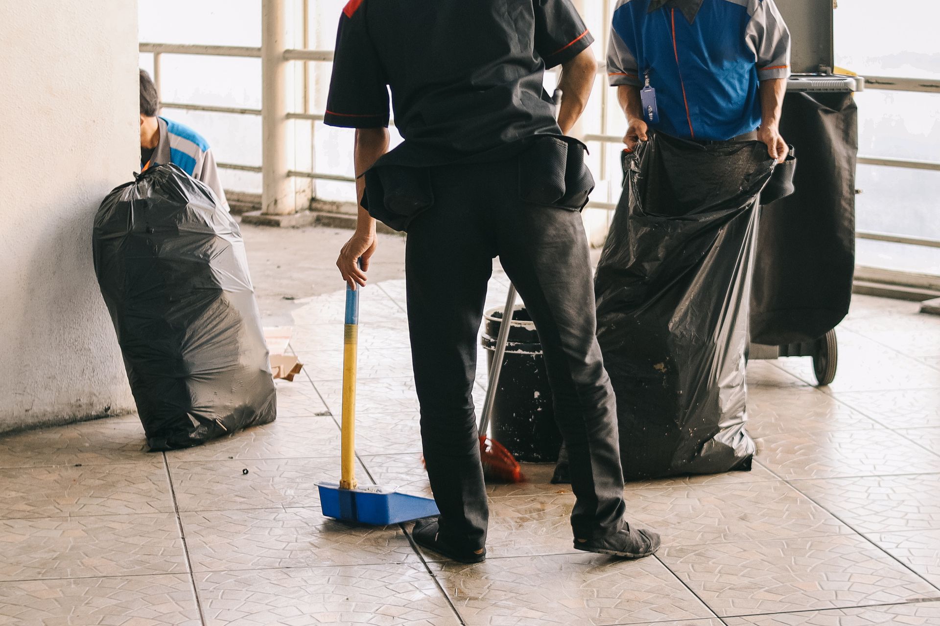 Two men are standing on a tiled floor holding trash bags and a broom.