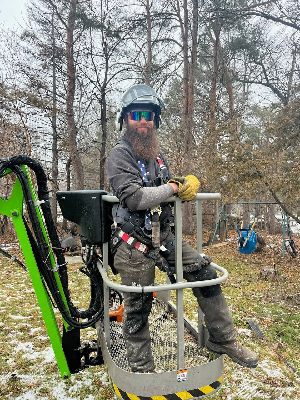 A man with a beard is sitting in a bucket on a lift in the woods.