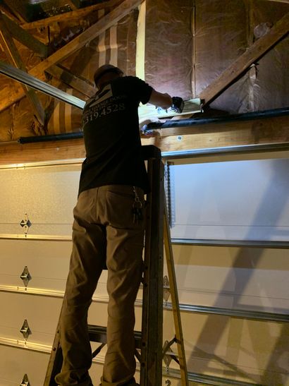 Person working overhead in a garage, using a tool on exposed wooden rafters above a closed garage door.