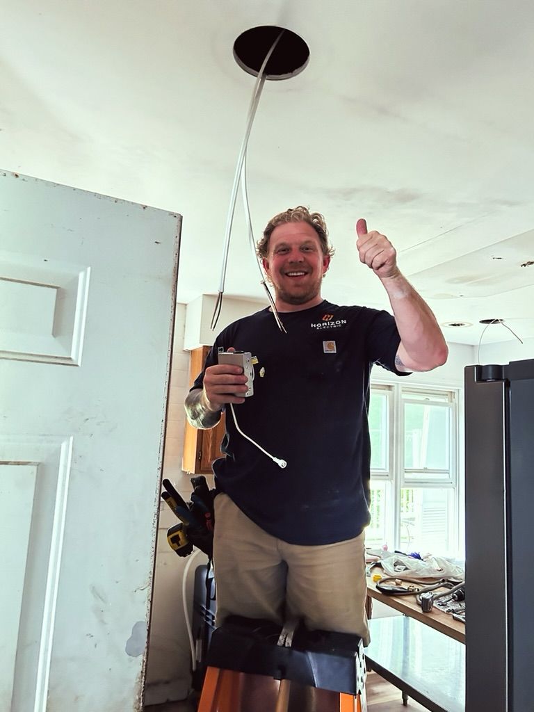 A person stands on a ladder in an unfinished room, giving a thumbs-up while holding wires from a ceiling light fixture.