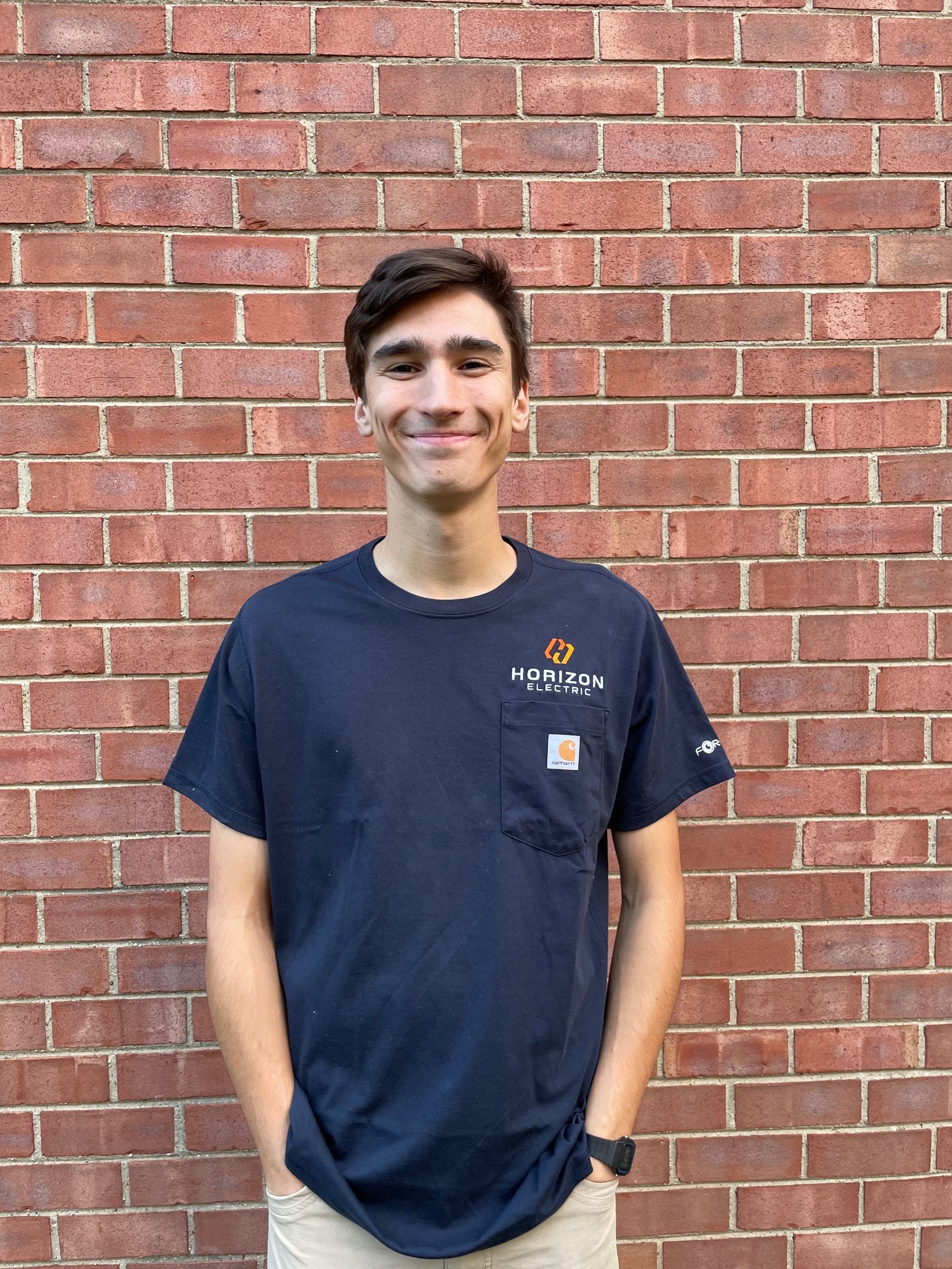 A smiling person wears a navy blue t-shirt with a pocket, standing in front of a red brick wall.