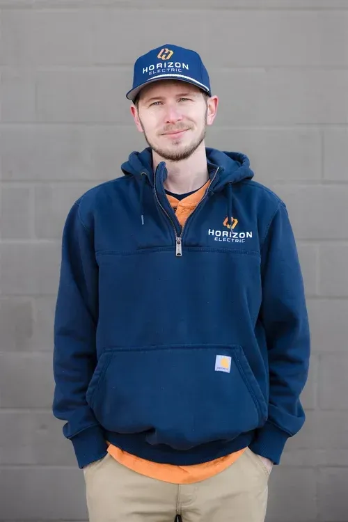 A person wearing a blue branded hoodie and hat with orange shirt underneath, posing against a grey block wall.