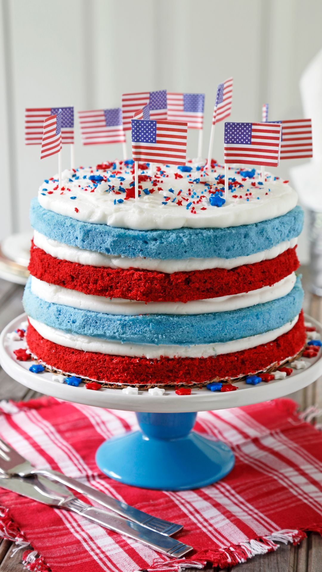 Red, white, and blue layered cake decorated with American flags, on a blue cake stand, and table setting.