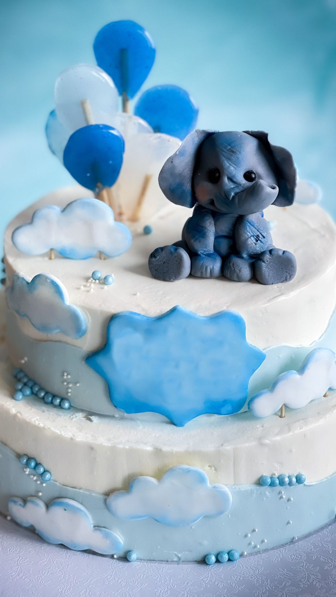 Two-tiered baby shower cake with blue fondant elephant, balloons, and clouds.