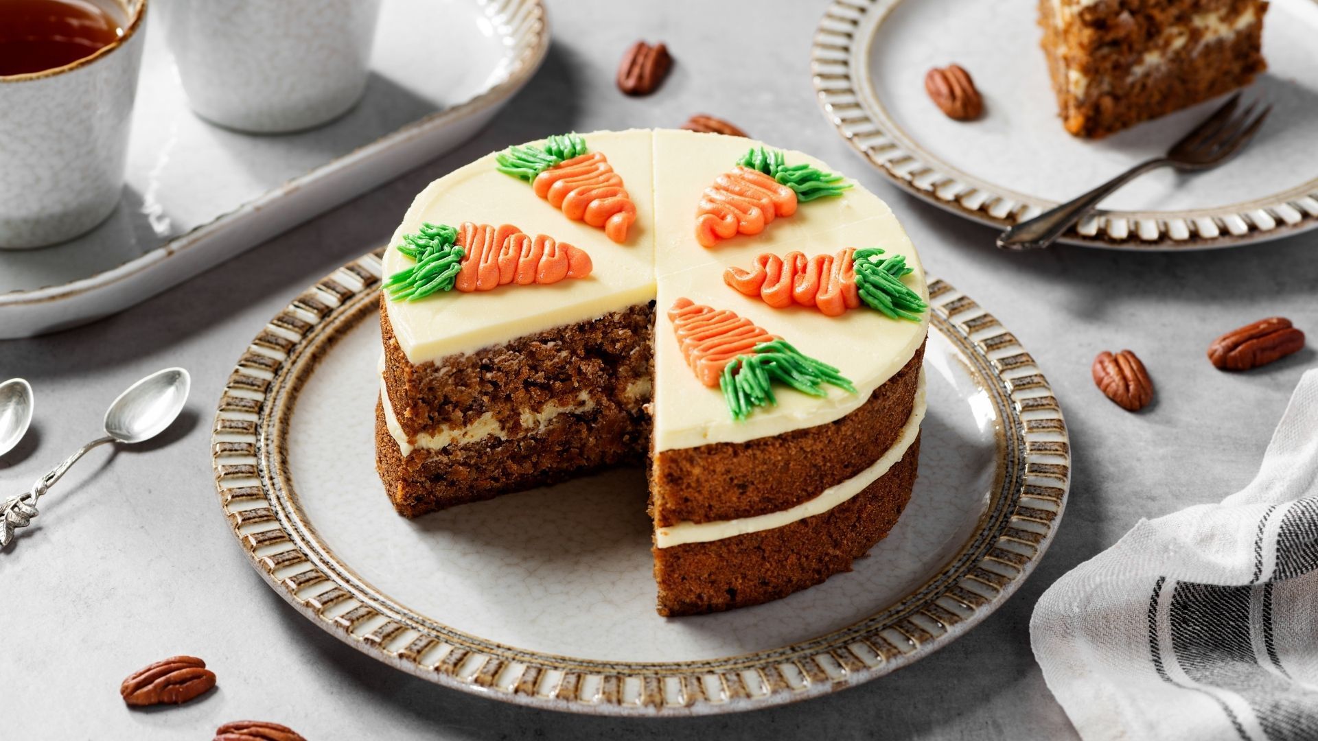 Carrot cake with frosting and carrot decorations on a plate with a slice missing, pecans, and tea.