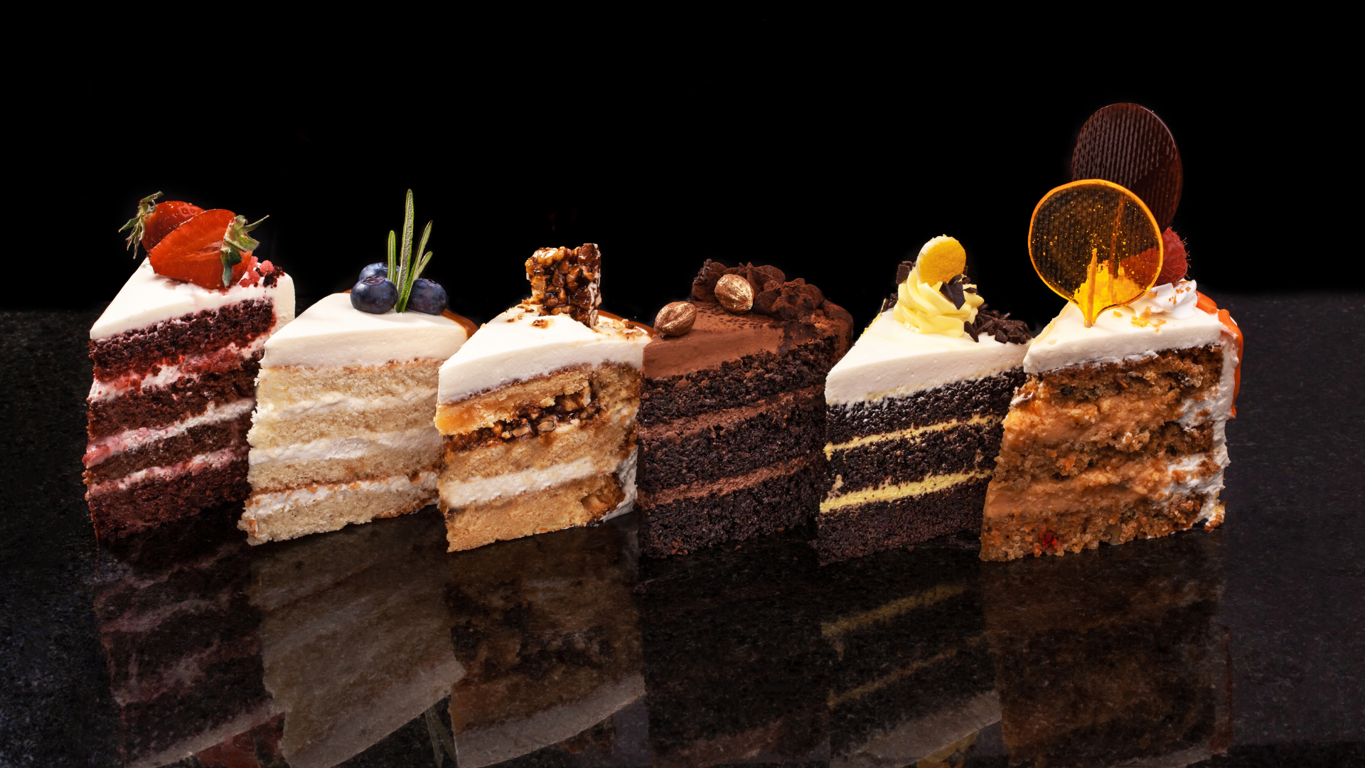 Slices of various cakes on a black surface, each with different flavors and toppings.