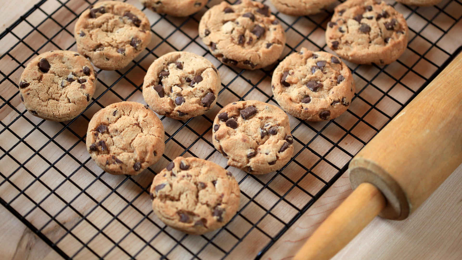 Chocolate chip cookies on a cooling rack next to a rolling pin, on a light-colored surface.