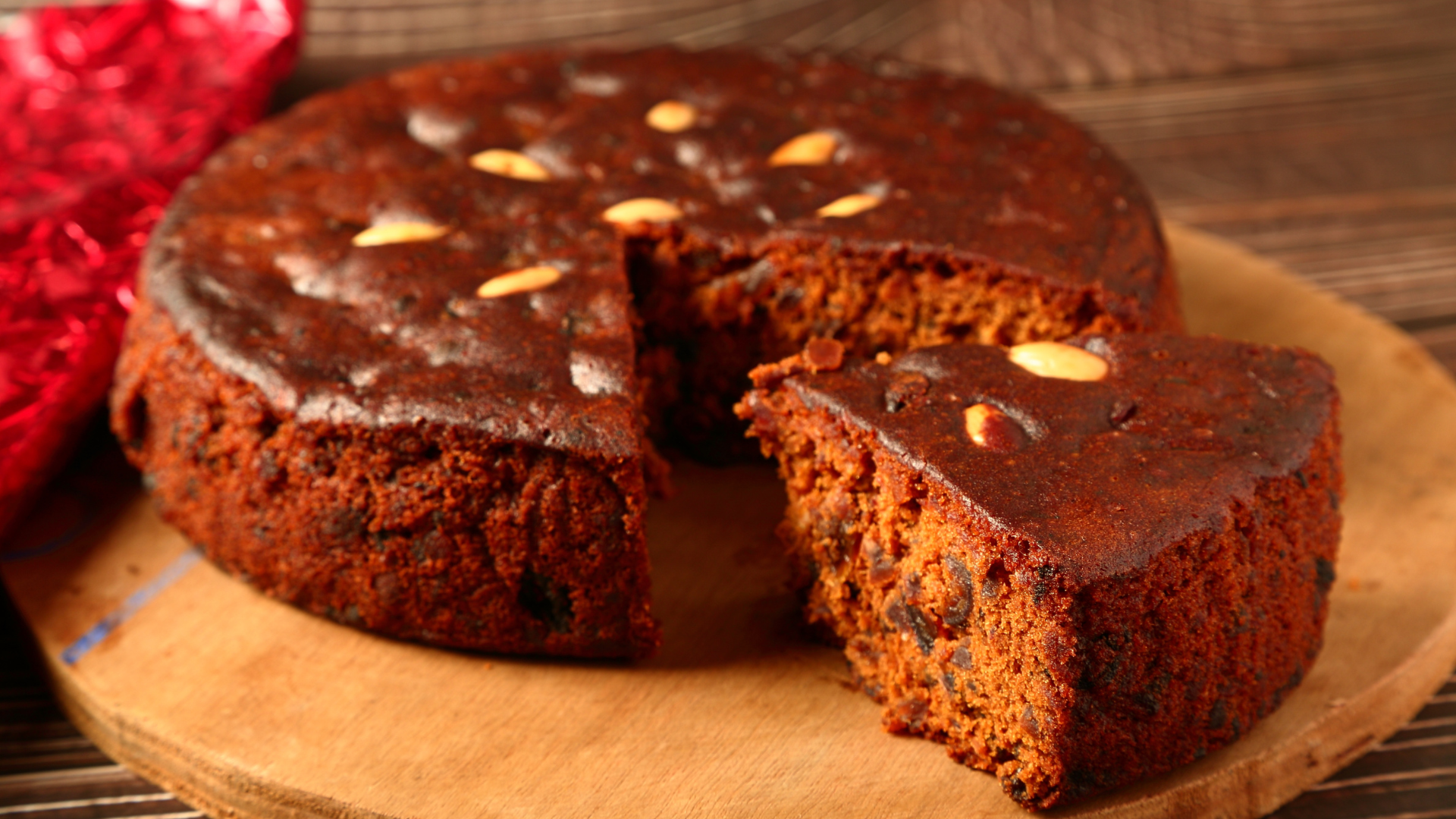 Round, dark fruitcake with a slice cut out, resting on a wooden board.