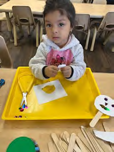 A little girl is sitting at a table making a snowman out of popsicle sticks.