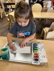 A little girl is sitting at a table painting with watercolors.