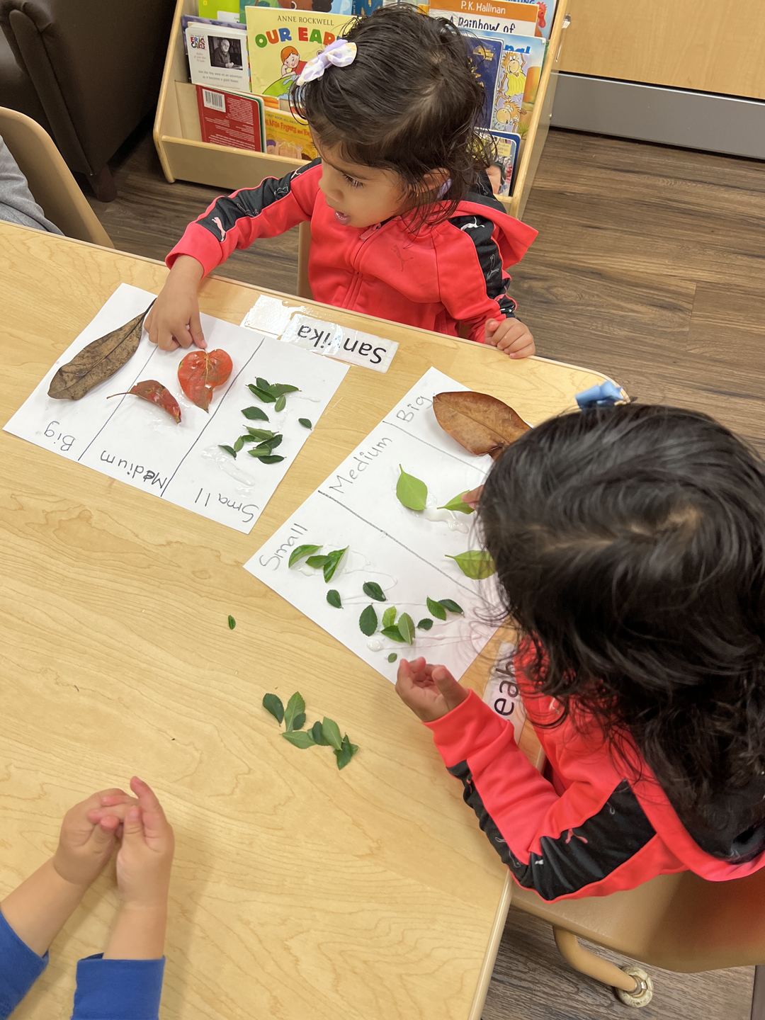 Two young girls are sitting at a table playing with leaves.