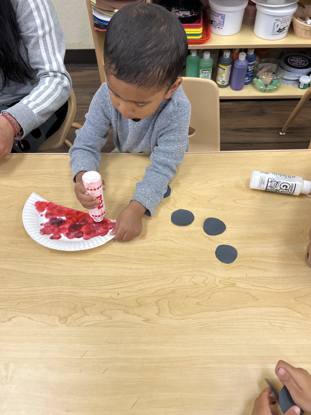 A little boy is sitting at a table making a paper plate watermelon.